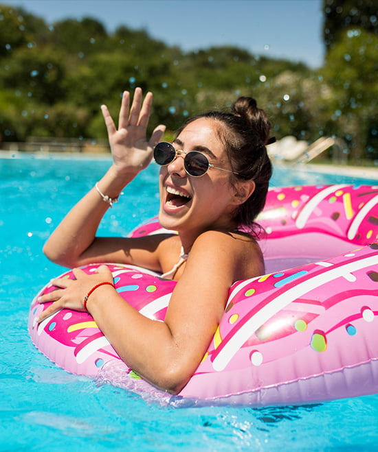 A smiling young woman in a pink donut float in the pool, wearing sunglasses and looking at the camera.